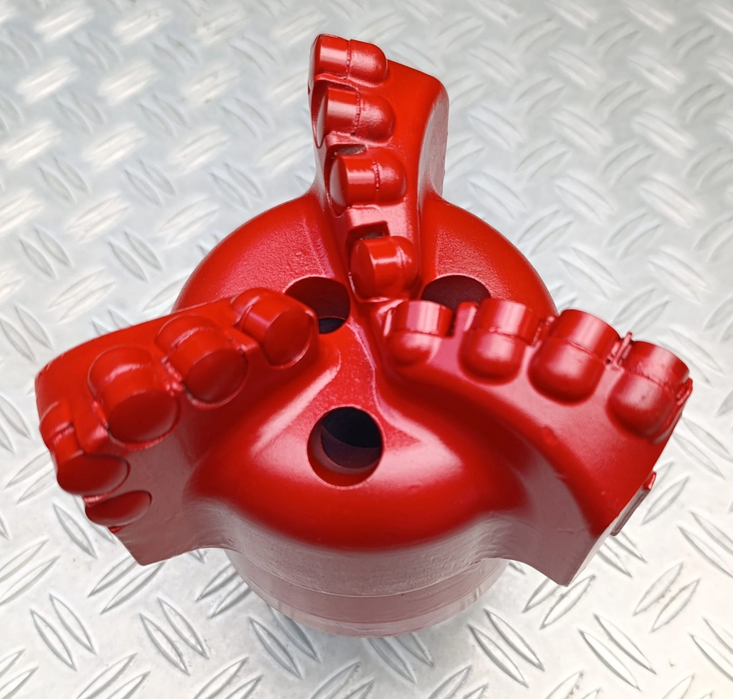H-Tools - Hochleistungs-Gesteinsbohrkronen - Ein leuchtend roter, mehrschneidiger PDC-Bohrer, möglicherweise ein 3- oder 5-schneidiges Modell, von vorne auf einer Metalloberfläche gezeigt.