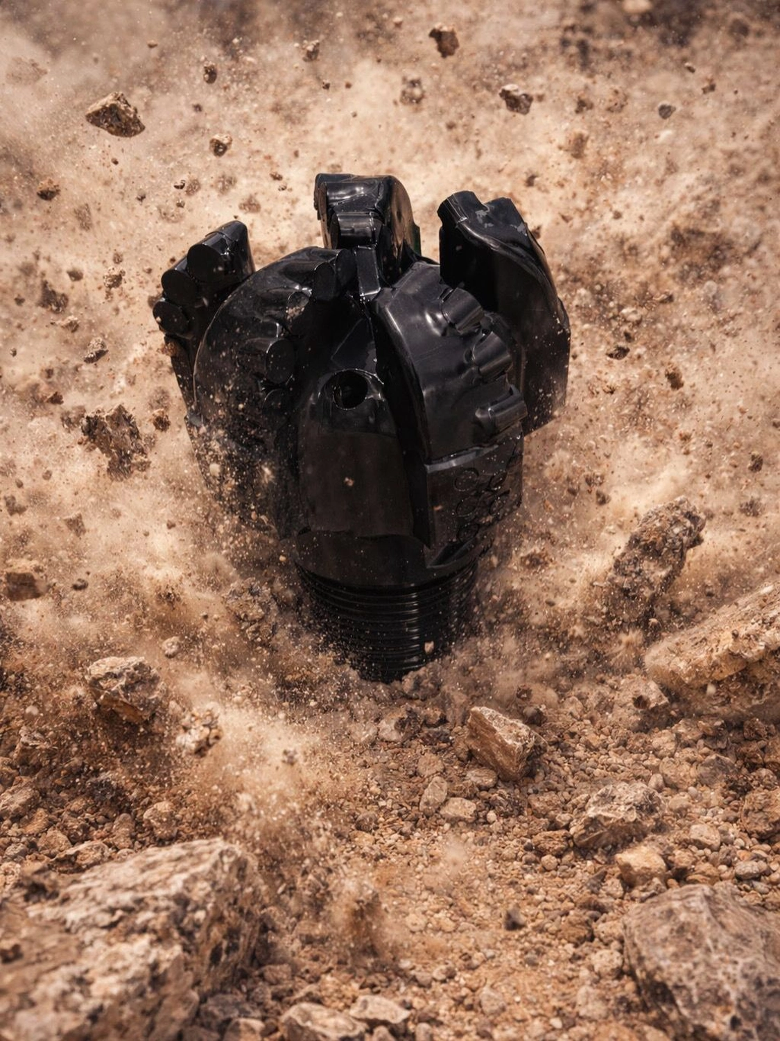 H-Tools - Hochleistungs-Gesteinsbohrkronen - Ein dunkelgrauer PDC-Bohrmeißel mit mehreren Schneiden, der teilweise in einer felsigen oder erdigen Bodenformation eingebettet ist.