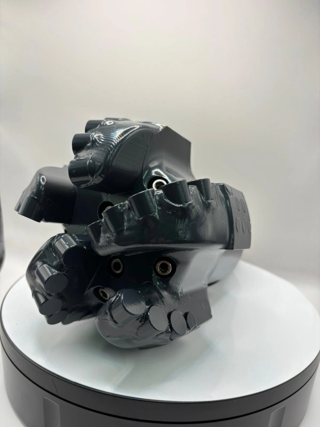 H-Tools - Hochleistungs-Gesteinsbohrkronen - Schräge Ansicht des großen, dunkelgrauen oder schwarzen Industriebohrkopfes mit mehreren Schneidmessern, der auf einem weißen, drehbaren Präsentationsständer ruht.